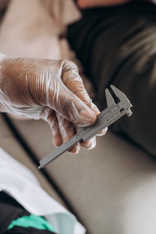 A gloved hand is holding a caliper, an instrument used for measuring dimensions. The background includes blurred fabric and possibly other indiscernible objects.