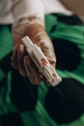 A gloved hand holds a sterile medical device, possibly for blood testing or sample collection, with a green and black background.