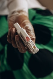A gloved hand holds a sterile medical device, possibly for blood testing or sample collection, with a green and black background.