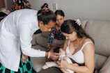A healthcare professional, dressed in white and wearing gloves, examines a newborn baby while the mother holds the baby on a sofa. A young girl is also present, sitting next to the mother and baby, observing attentively. There is a decorated Christmas tree in the background, indicating a festive atmosphere.
