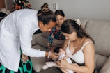 A healthcare professional, dressed in white and wearing gloves, examines a newborn baby while the mother holds the baby on a sofa. A young girl is also present, sitting next to the mother and baby, observing attentively. There is a decorated Christmas tree in the background, indicating a festive atmosphere.