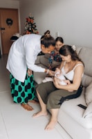 A woman in a white coat kneels beside a couch, attending to a baby held by another woman. A young girl watches the scene. A decorated Christmas tree is visible in the background, adding a festive touch to the cozy living room setting.