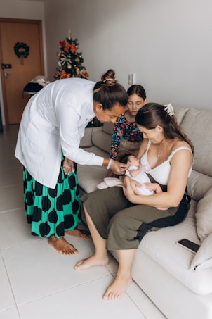 A woman in a white coat kneels beside a couch, attending to a baby held by another woman. A young girl watches the scene. A decorated Christmas tree is visible in the background, adding a festive touch to the cozy living room setting.
