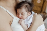 A newborn baby with soft dark hair sleeps peacefully against a caregiver's chest, dressed in a white outfit with small patterns. The scene exudes warmth and tenderness, with the caregiver's arm gently cradling the baby.