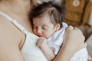 A newborn baby with soft dark hair sleeps peacefully against a caregiver's chest, dressed in a white outfit with small patterns. The scene exudes warmth and tenderness, with the caregiver's arm gently cradling the baby.