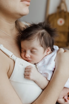 A sleeping baby with dark hair rests peacefully against an adult's chest, enveloped in a white garment. The baby's small hand is gently clenched, conveying a sense of security and warmth. The adult is wearing a white top, and a soft, earthy background with a hint of a brown bag adds to the serene atmosphere.