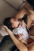 An infant, with dark hair, is being gently cradled in someone's arms. The baby is wearing a white onesie with small floral patterns, and the person's arm is supporting the baby's head and body. The scene conveys a sense of warmth and care.