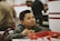 A young boy is sitting at a fast-food restaurant table, holding a hamburger and looking content. He is wearing a gray hoodie with a blue hood visible around the collar. In front of him is a red tray with fast-food packaging. The background shows blurred figures and a casual dining setting.