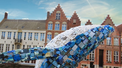 A large sculpture resembling a whale is composed entirely of blue and white plastics. The background features historic European-style brick buildings with gabled roofs. The sky is clear with a few clouds.