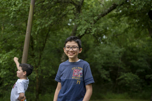 A young boy wearing a navy blue shirt with a print, standing in a lush green forest area. He is smiling directly at the camera. Another boy is in the background, wearing a white shirt, looking off to the side with one arm raised.