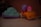 Two toy vehicles are placed on a smooth, light-colored surface. The left toy is shaped like a blue frog with purple wheels, showing signs of wear and some dirt. The right toy is a small orange car with a cartoonish character wearing a pink helmet seated on top. The character's head is white with minimal features.