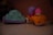 Two toy vehicles are placed on a smooth, light-colored surface. The left toy is shaped like a blue frog with purple wheels, showing signs of wear and some dirt. The right toy is a small orange car with a cartoonish character wearing a pink helmet seated on top. The character's head is white with minimal features.