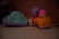 Two toy vehicles are placed on a smooth, light-colored surface. The left toy is shaped like a blue frog with purple wheels, showing signs of wear and some dirt. The right toy is a small orange car with a cartoonish character wearing a pink helmet seated on top. The character's head is white with minimal features.