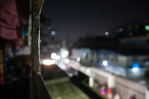 A metal railing is in focus in the foreground, with an urban night scene blurred in the background. The scene includes out-of-focus city lights and indistinct structures.