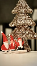 A decorative golden Christmas tree with small Santa figurines in red and white outfits is placed on a white surface. The tree is adorned with shiny tinsel, giving it a festive appearance.