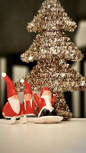 A decorative golden Christmas tree with small Santa figurines in red and white outfits is placed on a white surface. The tree is adorned with shiny tinsel, giving it a festive appearance.