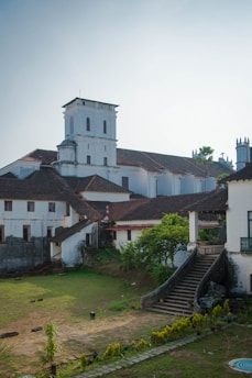 A historical building characterized by its colonial architecture, featuring white walls and sloping tiled roofs. The structure includes a tall tower with small windows and adjacent smaller buildings. A staircase leads up to the main entrance, surrounded by greenery and a spacious courtyard.