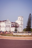 A large, white colonial-era building with architectural details surrounded by greenery. A sign in front reads 'Archaeological Survey of India'. A group of people are gathered near the entrance. Tall trees can be seen on either side of the building.