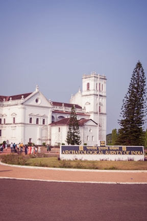 A large, white colonial-era building with architectural details surrounded by greenery. A sign in front reads 'Archaeological Survey of India'. A group of people are gathered near the entrance. Tall trees can be seen on either side of the building.