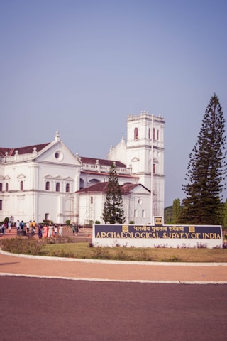 A large, white colonial-era building with architectural details surrounded by greenery. A sign in front reads 'Archaeological Survey of India'. A group of people are gathered near the entrance. Tall trees can be seen on either side of the building.