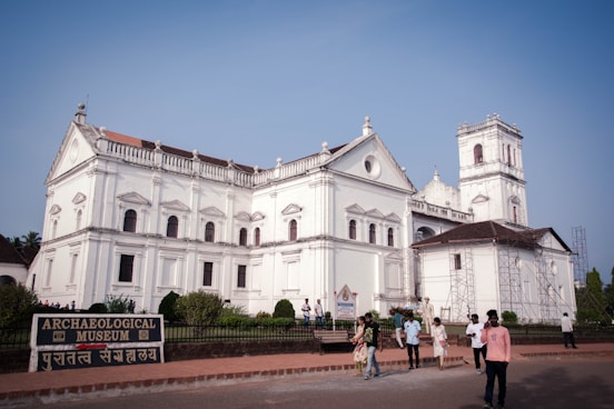A large historical white building with classical architecture, featuring ornate detailing and a prominent facade, likely a church or museum. People walk around the entrance area, and there is a sign reading 'Archaeological Museum' in English and another language. The sky is clear and blue, enhancing the building's grandeur.