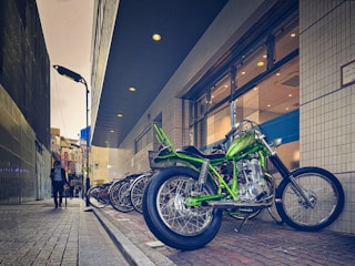 A vibrant green motorcycle is parked on a brick sidewalk next to a row of bicycles. The setting is an urban alleyway beside a modern building with glass windows and patterned walls. A person in business attire walks down the sidewalk, holding a phone. The scene is illuminated by streetlights and the ambient light of the overcast sky.