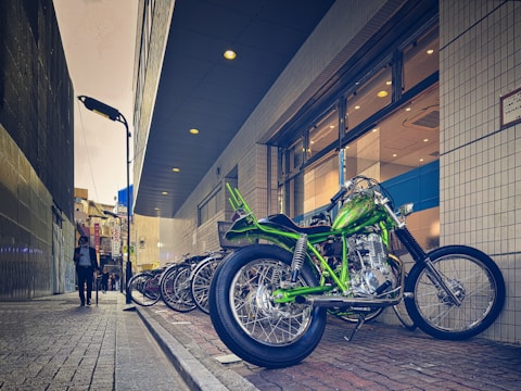 A vibrant green motorcycle is parked on a brick sidewalk next to a row of bicycles. The setting is an urban alleyway beside a modern building with glass windows and patterned walls. A person in business attire walks down the sidewalk, holding a phone. The scene is illuminated by streetlights and the ambient light of the overcast sky.