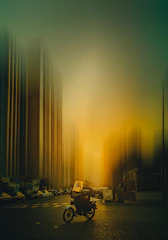 Motorcyclist in yellow and soft gold tones riding through a narrow urban street with hints of light blue in the sky.