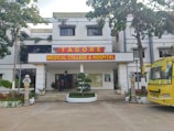 The image depicts the main entrance of a medical college and hospital, which has the name 'Tagore Medical College & Hospital' prominently displayed above the doorway. The building is a white multi-story structure with a clean, organized appearance, flanked by some trees and greenery. A yellow college bus with the name 'Tagore College Bus' is parked on the right side. A few potted plants and decorative pillars are present around the entrance.