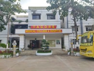 The image depicts the main entrance of a medical college and hospital, which has the name 'Tagore Medical College & Hospital' prominently displayed above the doorway. The building is a white multi-story structure with a clean, organized appearance, flanked by some trees and greenery. A yellow college bus with the name 'Tagore College Bus' is parked on the right side. A few potted plants and decorative pillars are present around the entrance.
