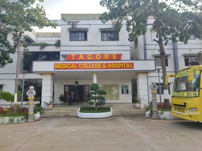 The image depicts the main entrance of a medical college and hospital, which has the name 'Tagore Medical College & Hospital' prominently displayed above the doorway. The building is a white multi-story structure with a clean, organized appearance, flanked by some trees and greenery. A yellow college bus with the name 'Tagore College Bus' is parked on the right side. A few potted plants and decorative pillars are present around the entrance.