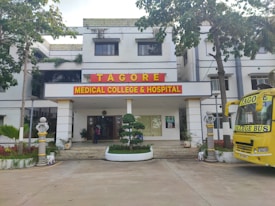 The image depicts the main entrance of a medical college and hospital, which has the name 'Tagore Medical College & Hospital' prominently displayed above the doorway. The building is a white multi-story structure with a clean, organized appearance, flanked by some trees and greenery. A yellow college bus with the name 'Tagore College Bus' is parked on the right side. A few potted plants and decorative pillars are present around the entrance.