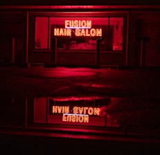 A neon sign glowing in bright red with the words 'Fusion Hair Salon' is prominently displayed. The sign is reflected on a wet surface below, likely a sidewalk or street, creating a mirror image. The surrounding environment is dim, with dark brick walls and window frames, emphasizing the vibrant neon glow.