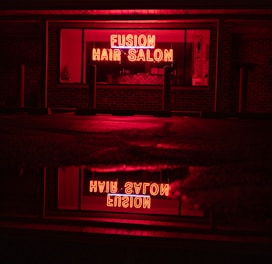 A neon sign glowing in bright red with the words 'Fusion Hair Salon' is prominently displayed. The sign is reflected on a wet surface below, likely a sidewalk or street, creating a mirror image. The surrounding environment is dim, with dark brick walls and window frames, emphasizing the vibrant neon glow.
