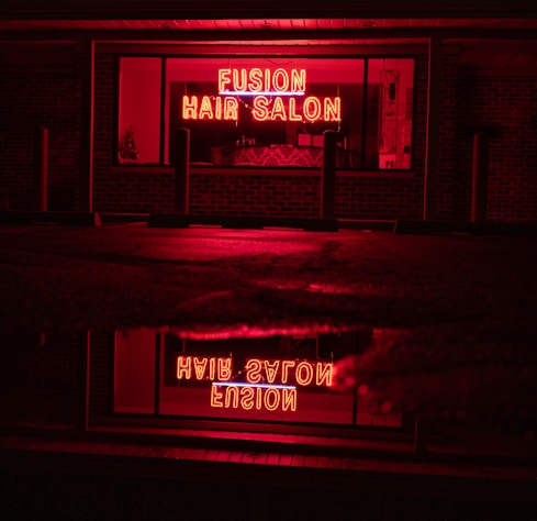 A neon sign glowing in bright red with the words 'Fusion Hair Salon' is prominently displayed. The sign is reflected on a wet surface below, likely a sidewalk or street, creating a mirror image. The surrounding environment is dim, with dark brick walls and window frames, emphasizing the vibrant neon glow.