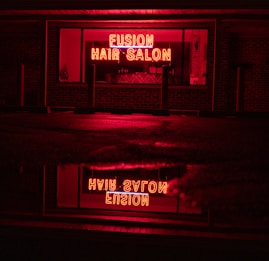 A neon sign glowing in bright red with the words 'Fusion Hair Salon' is prominently displayed. The sign is reflected on a wet surface below, likely a sidewalk or street, creating a mirror image. The surrounding environment is dim, with dark brick walls and window frames, emphasizing the vibrant neon glow.