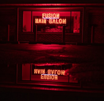 A neon sign glowing in bright red with the words 'Fusion Hair Salon' is prominently displayed. The sign is reflected on a wet surface below, likely a sidewalk or street, creating a mirror image. The surrounding environment is dim, with dark brick walls and window frames, emphasizing the vibrant neon glow.