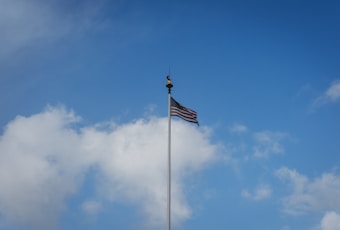 A flagpole with an American flag waving against a backdrop of a bright blue sky with scattered white clouds. The flag is positioned toward the top of the image, and the flagpole is centered.