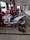 A white and blue motorcycle is positioned on a hydraulic lift within a garage, surrounded by various tools on the floor. The garage has several other motorcycles being serviced. The space is lit by natural light coming through open doors, and the floor is tiled.
