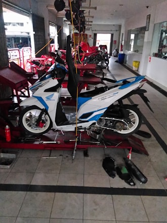 A white and blue motorcycle is positioned on a hydraulic lift within a garage, surrounded by various tools on the floor. The garage has several other motorcycles being serviced. The space is lit by natural light coming through open doors, and the floor is tiled.