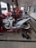 A white and blue motorcycle is positioned on a hydraulic lift within a garage, surrounded by various tools on the floor. The garage has several other motorcycles being serviced. The space is lit by natural light coming through open doors, and the floor is tiled.