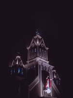 A religious building, possibly a church, illuminated against a dark night sky. Stained glass windows are visible on the structure, and a statue of Jesus Christ wearing a red robe stands prominently on the front facade.