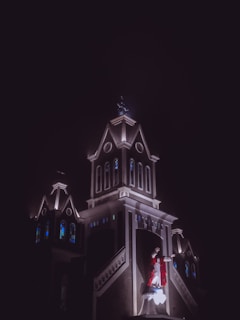 A religious building, possibly a church, illuminated against a dark night sky. Stained glass windows are visible on the structure, and a statue of Jesus Christ wearing a red robe stands prominently on the front facade.
