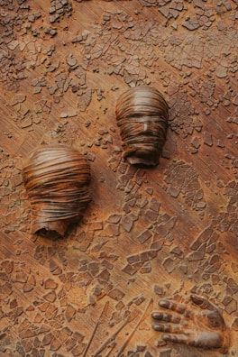 The artwork features two stylized, textured heads wrapped in cloth and a sculpted hand against a cracked, earthy background. The textures give a sense of decay or transformation.
