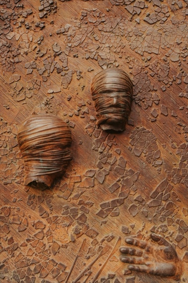The artwork features two stylized, textured heads wrapped in cloth and a sculpted hand against a cracked, earthy background. The textures give a sense of decay or transformation.