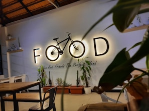 A cozy indoor cafe setting featuring a decorative wall with the word 'FOOD' creatively incorporating a bicycle as the 'O'. Below, potted plants and minimalistic wooden tables and chairs contribute to a warm, inviting atmosphere.