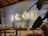 A cozy indoor cafe setting featuring a decorative wall with the word 'FOOD' creatively incorporating a bicycle as the 'O'. Below, potted plants and minimalistic wooden tables and chairs contribute to a warm, inviting atmosphere.