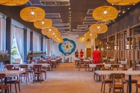A spacious and warmly lit dining area with wooden tables and chairs arranged neatly. Large wicker pendant lights hang from the ceiling, casting a cozy glow. White drapes cover the tall windows on the left side, adding an elegant touch. The far end of the room features a large decorative wall piece with a blue and green floral design. Two people in red shirts are standing near this area, suggesting a restaurant setting.