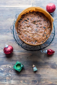 A fruitcake is placed on a wire cooling rack on a wooden table. It is surrounded by red Christmas ornaments, a small green decorative item, and a miniature figure of Santa Claus.