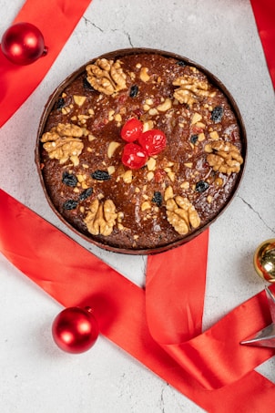 A richly decorated fruitcake is topped with walnuts, red cherries, and mixed nuts, surrounded by a vibrant red ribbon and Christmas ornaments on a light-colored surface.